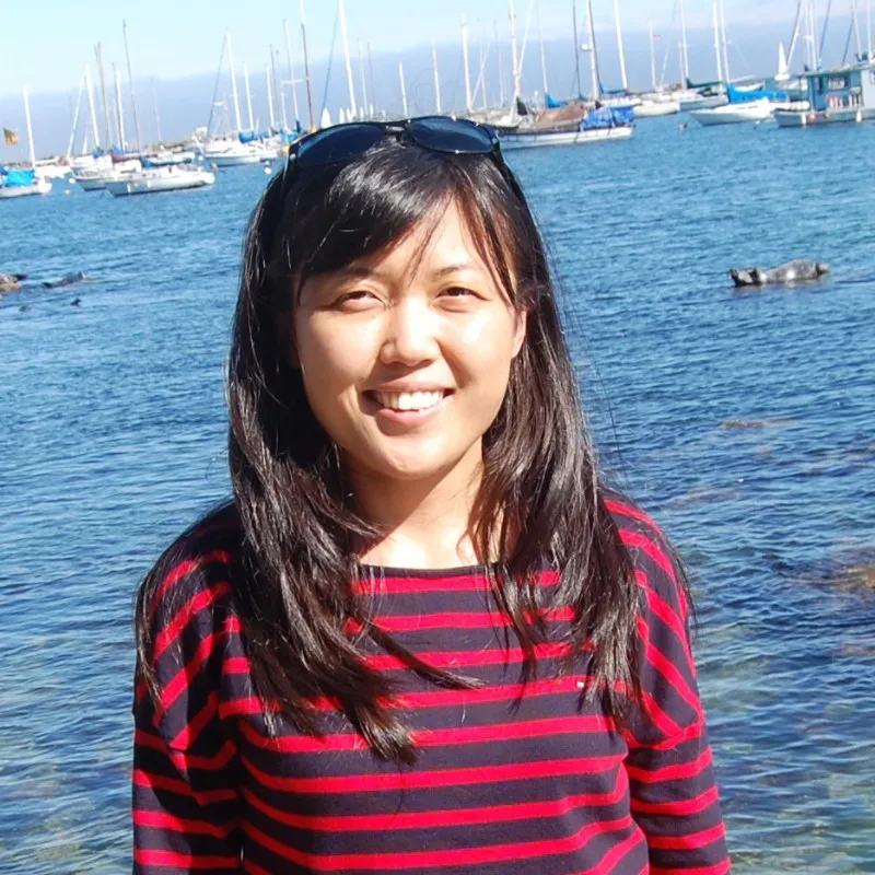 Yujia Zhao smiling to camera, wearing a red and navy striped top with sunglasses pushed up over dark hair, in front of a harbour of moored sailboats and clear blue water.